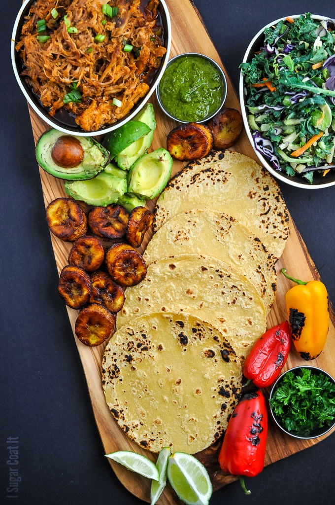 a wooden board with all the fixings for tacos including plantains, avocado grilled vegetables, taco shells and pulled pork | i sugar coat it