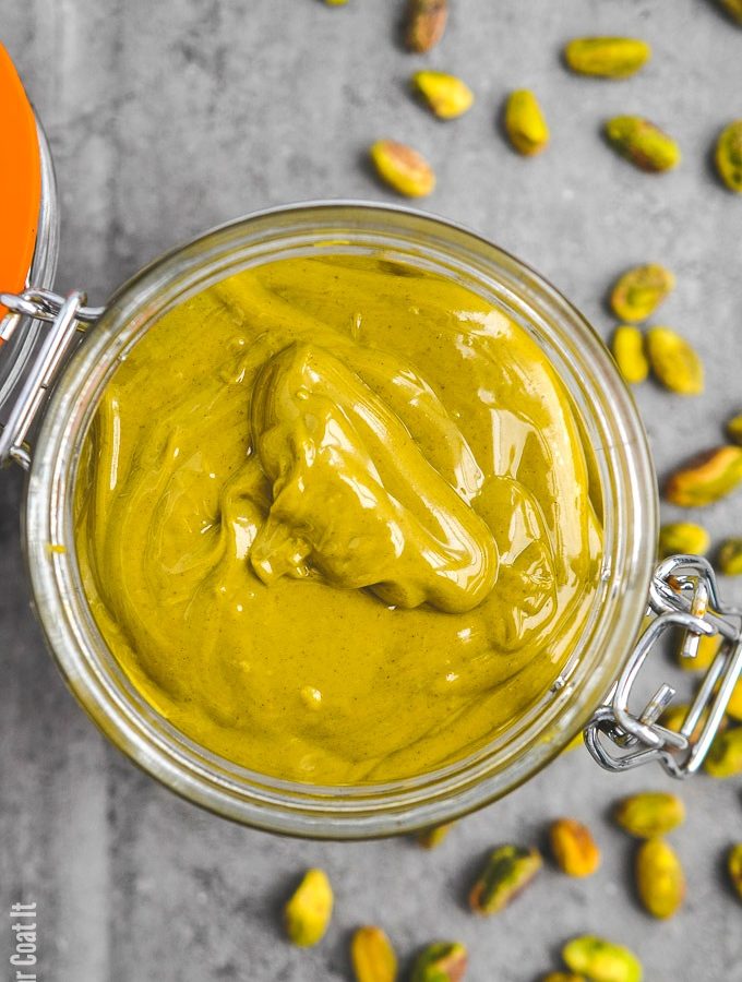 Pistachio paste in a jar with pistachios scattered around. | I Sugar Coat It