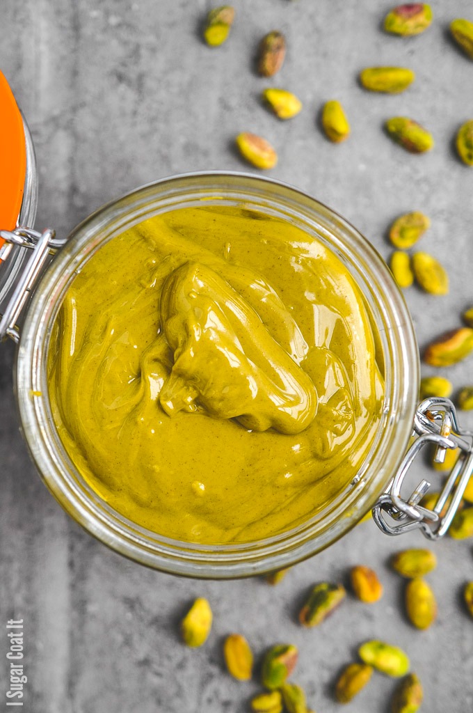 Pistachio paste in a jar with pistachios scattered around. | I Sugar Coat It